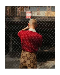 Man with short blonde hair seen from behind wearing a patterned red polo shirt and tan trousers standing behind a chain-link fence.