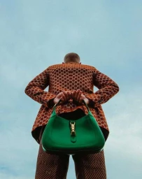 Low-angle shot of a man wearing a patterned rust-orange blazer holding a green leather handbag against a pale blue sky.