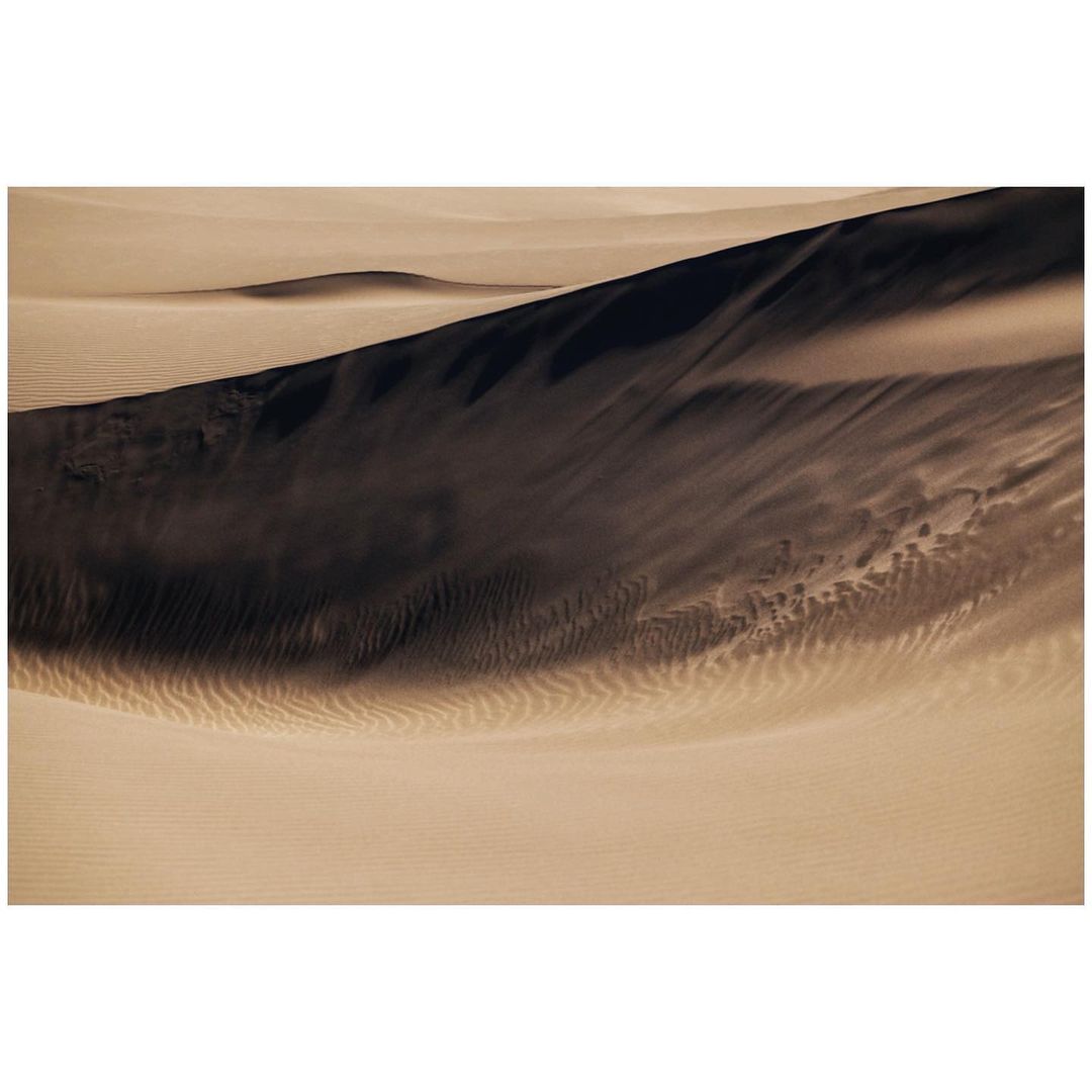 Dramatic, abstract landscape of sand dunes with rippled textures, and strong contrast between light and shadow in an arid environment.