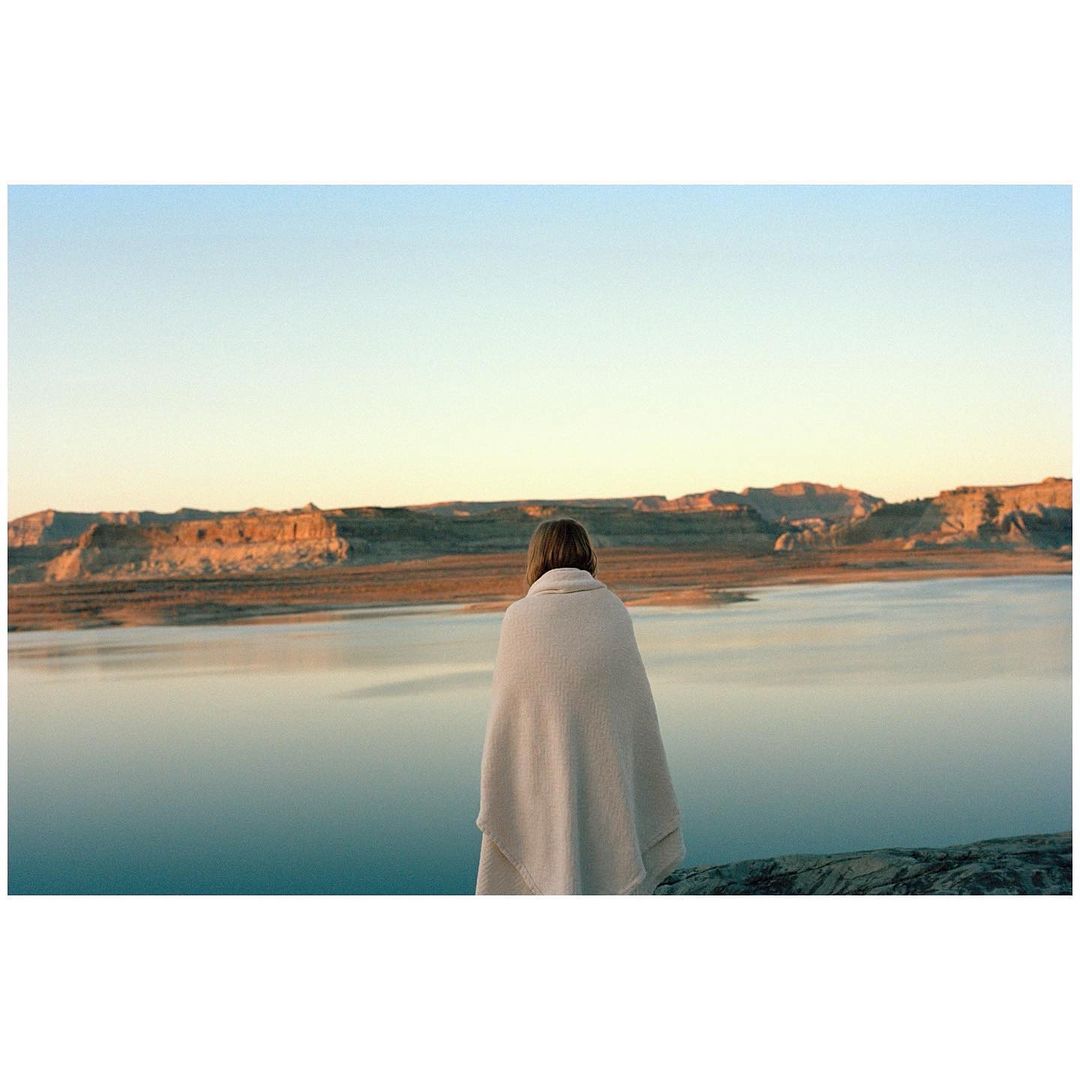 Woman wrapped in a blanket gazes at a lake surrounded by rocky hills during sunset. The scene exudes serenity and natural beauty.