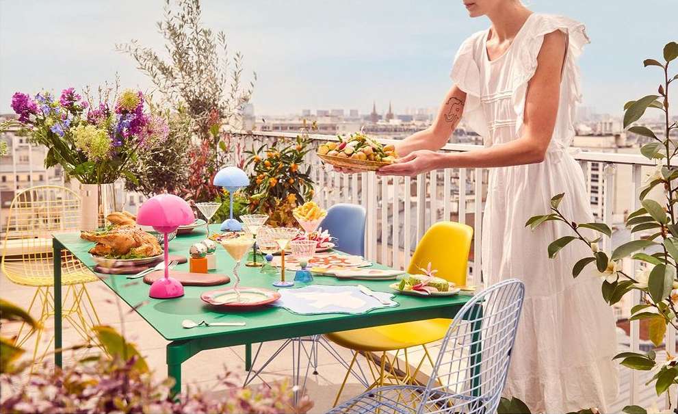 Woman in white dress serves food at outdoor rooftop table with bright chairs, colorful lamps, and cityscape view.