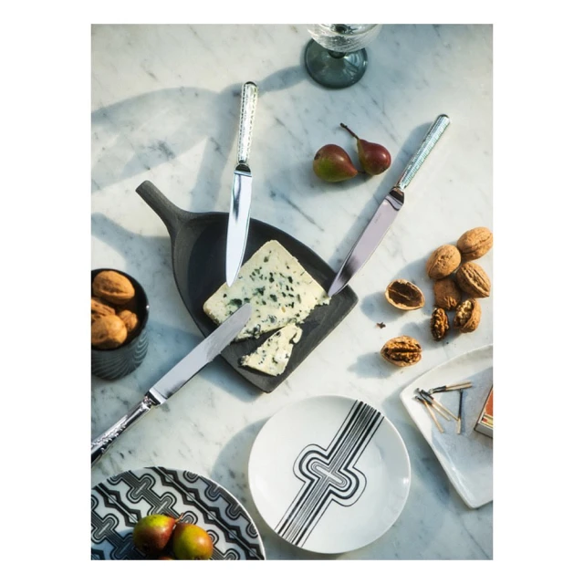 Overhead shot of a cheese board with blue cheese, pears, walnuts, ornate knives, decorative plates and glasses on a marble table.