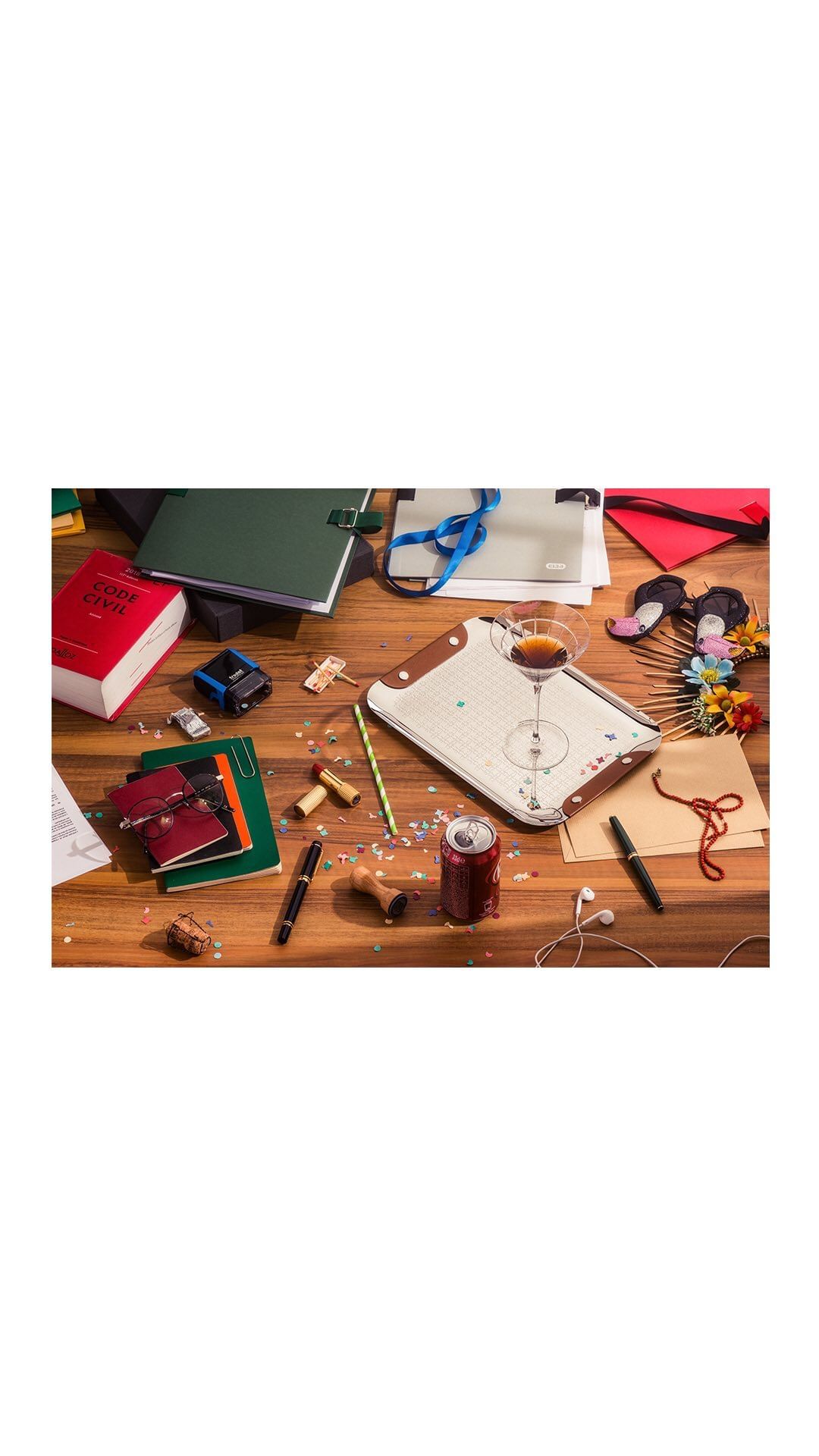 Overhead studio shot of a wood desk covered with a cocktail glass, notebooks, pens, a soda can, corks, headphones, and various office supplies.