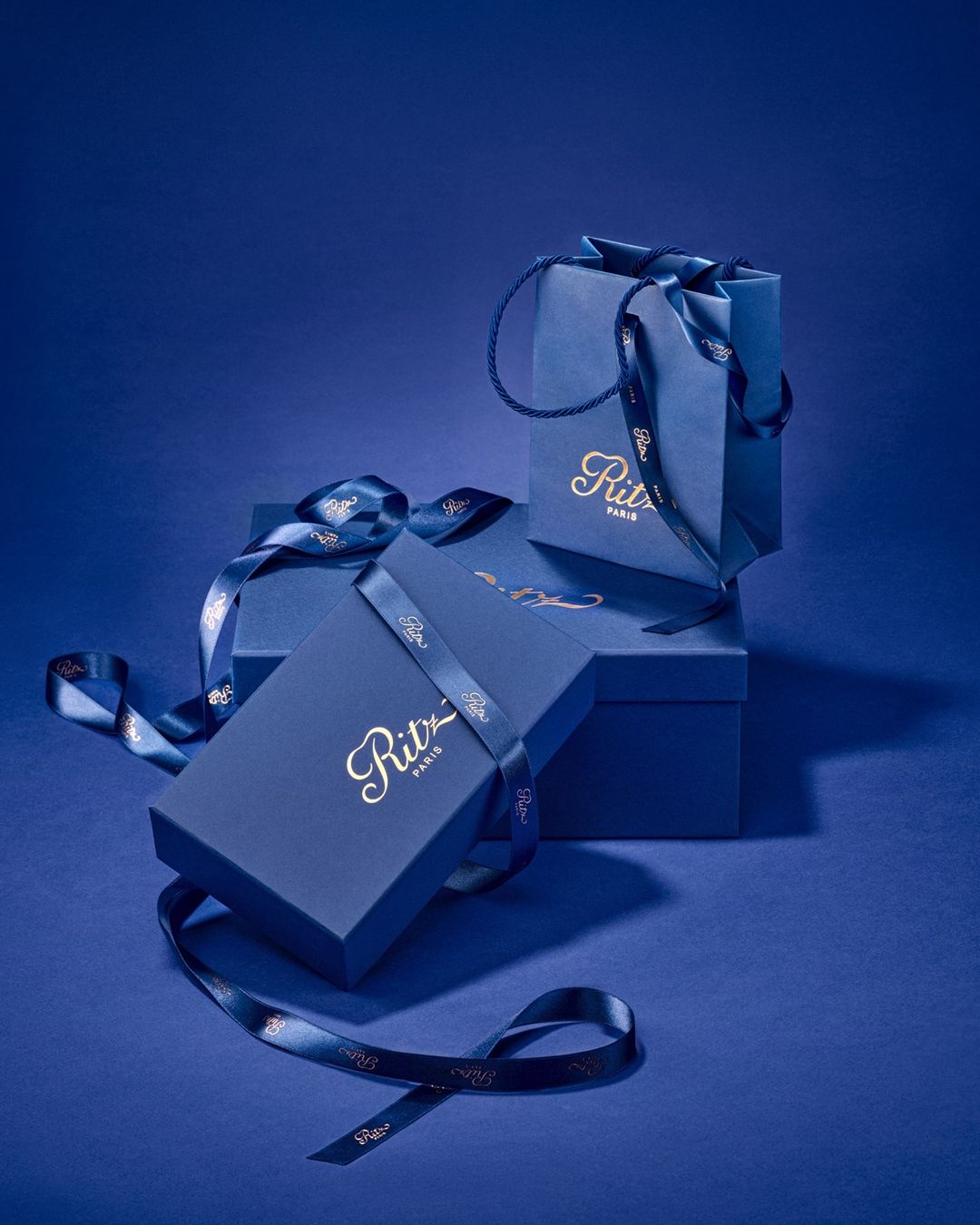 Elegant presentation of a gift box, shopping bag, and ribbon in shades of blue with gold lettering against a solid blue background.