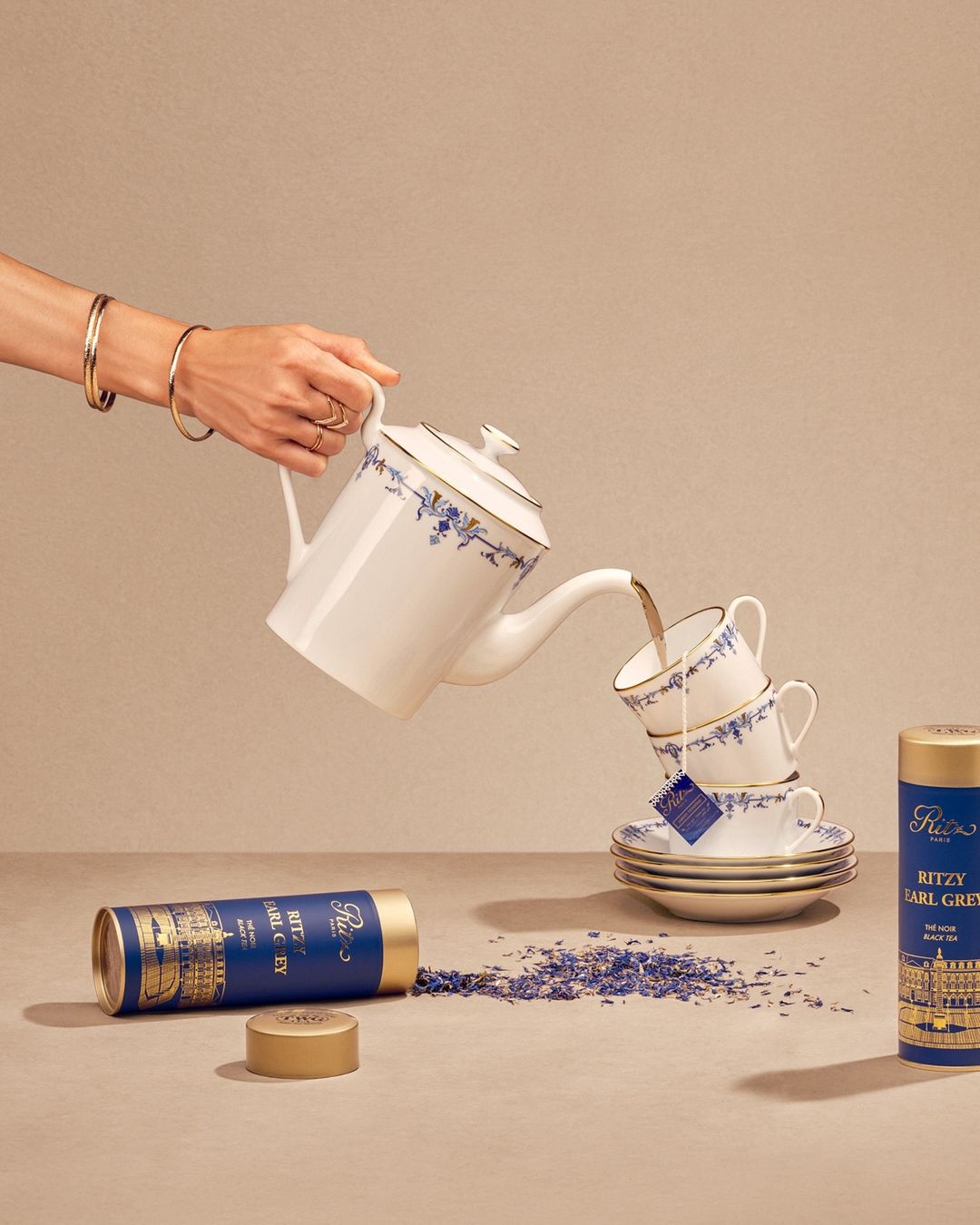 Tea is poured into cups from a white teapot with gold trim. Tea tins are stacked nearby on a beige surface with floral patterns.