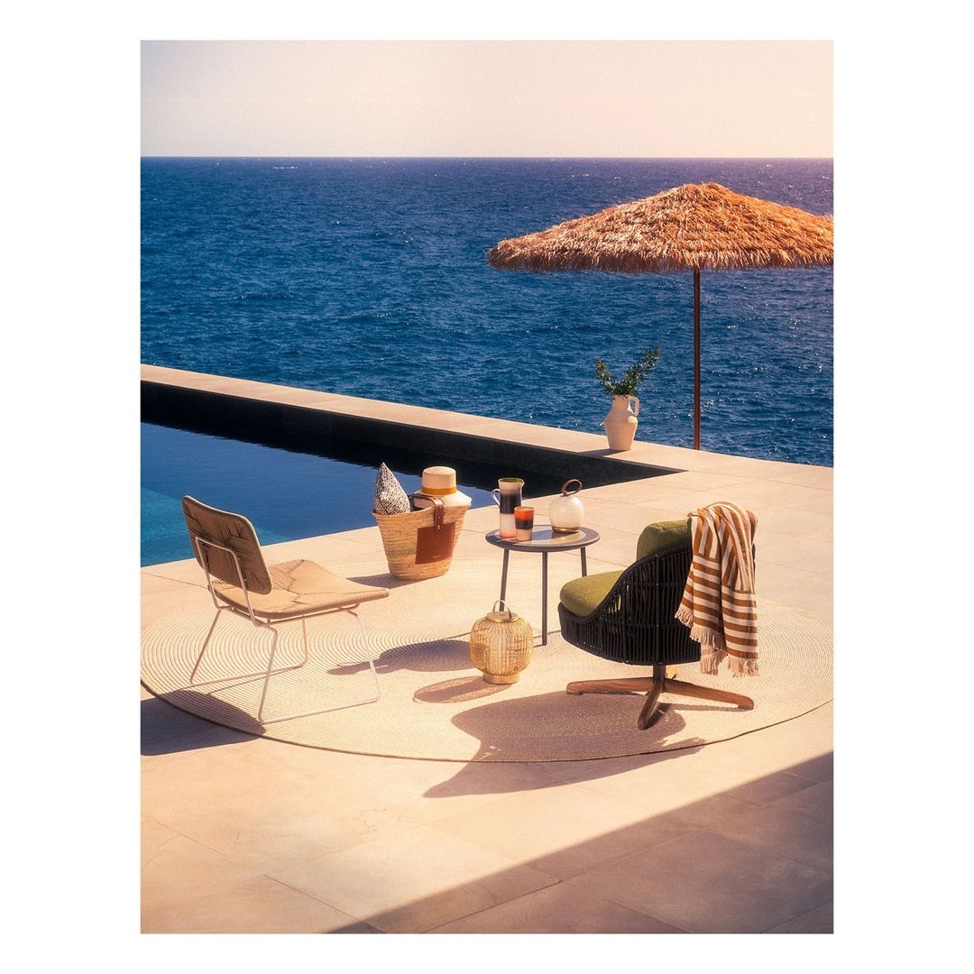 Outdoor seating arrangement on a patio overlooking the ocean with chairs, table, umbrella, and straw bag in warm, sunny daylight.