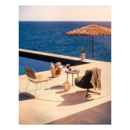Outdoor seating arrangement on a patio overlooking the ocean with chairs, table, umbrella, and straw bag in warm, sunny daylight.