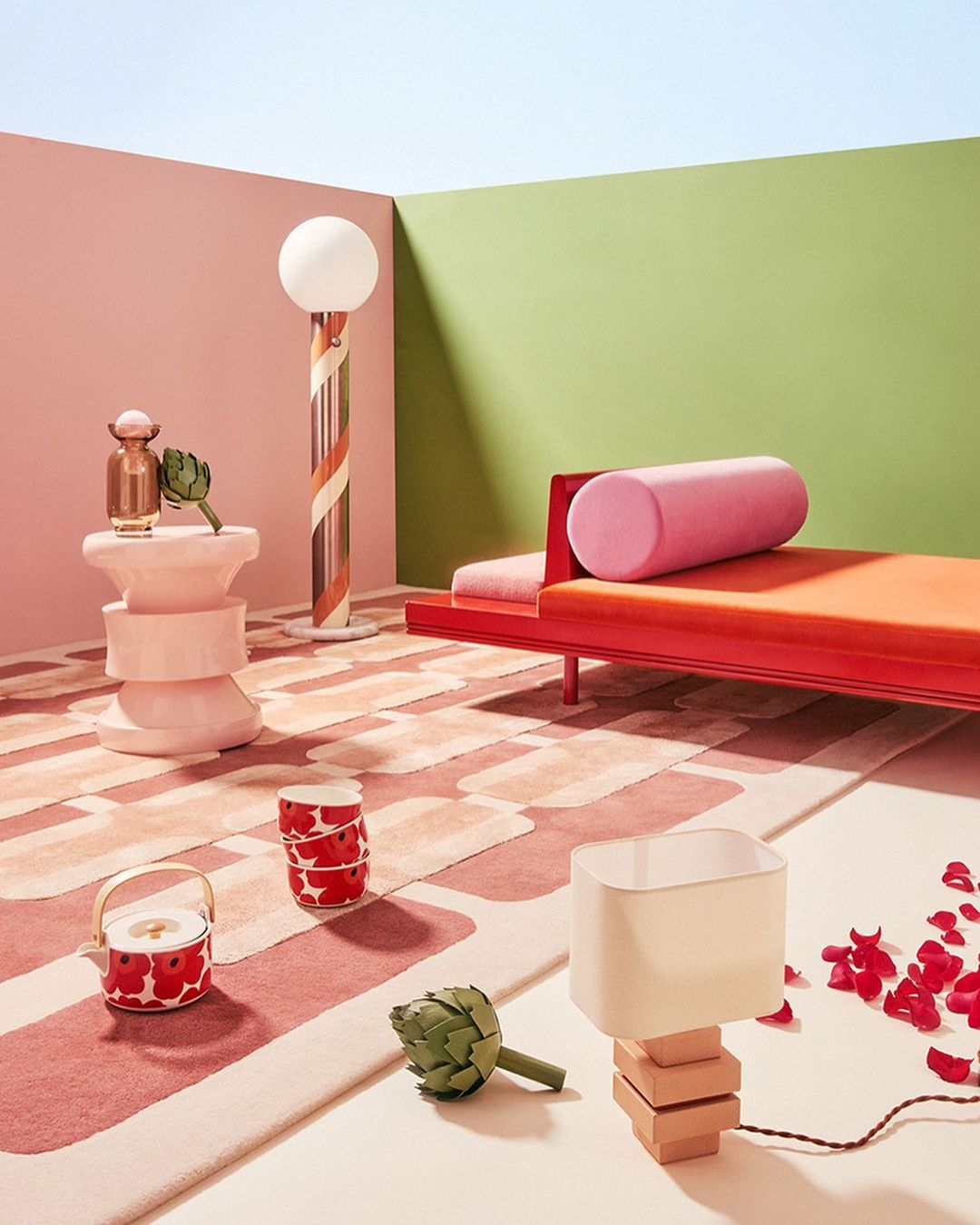 Interior scene with a coral chaise lounge, unique lamps, decorative vase, artichokes, tea set on a patterned rug in a pastel room.