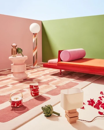 Interior scene with a coral chaise lounge, unique lamps, decorative vase, artichokes, tea set on a patterned rug in a pastel room.
