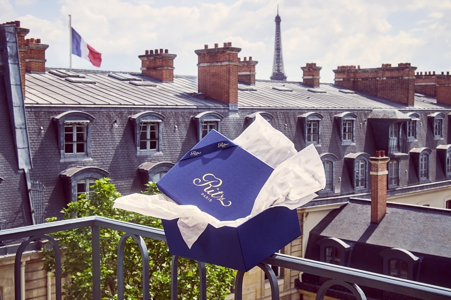 A box from the Ritz Paris sits on a balcony railing with a view of Paris rooftops and the Eiffel Tower in the background.