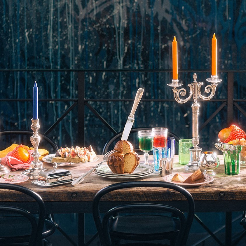 Still life scene with a wooden table set with cake, fruit, and colorful drinks, illuminated by candles in silver candlesticks against a dark backdrop.