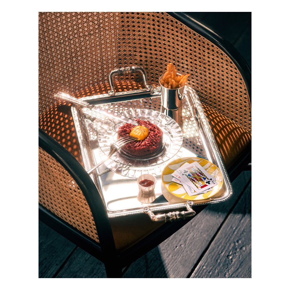 Elevated shot of silver serving tray with steak tartare, french fries, cocktail, and playing cards, set on rattan chair on wooden surface.