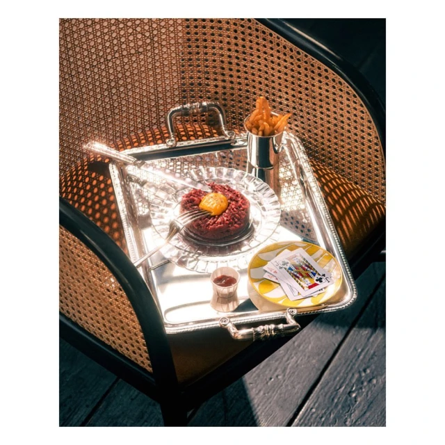 Elevated shot of silver serving tray with steak tartare, french fries, cocktail, and playing cards, set on rattan chair on wooden surface.