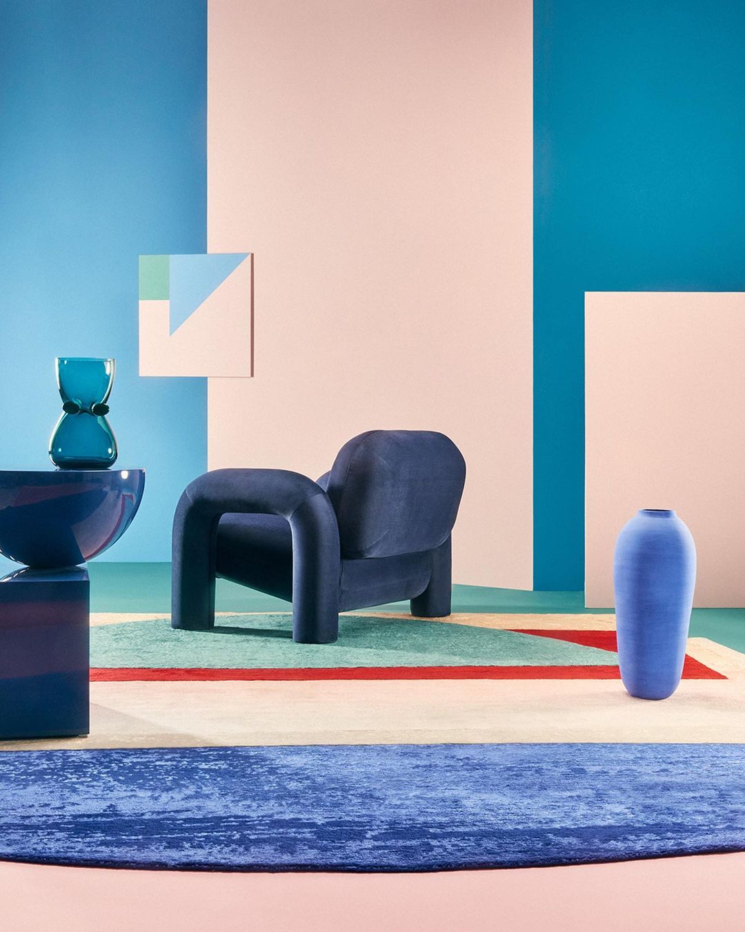 Modern interior design featuring a blue armchair, vases, and a geometric rug against a backdrop of pastel color blocks.