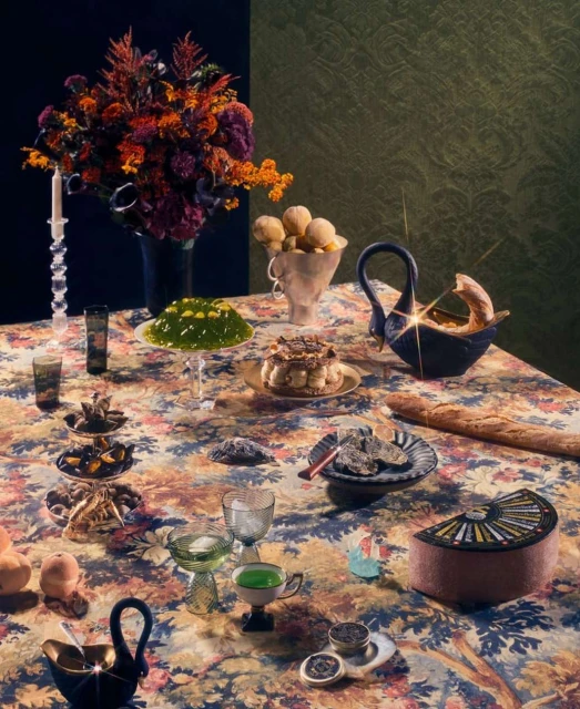Still life featuring floral tablecloth, an assortment of desserts, fruits, and vintage glassware arranged on a table against a dark backdrop.