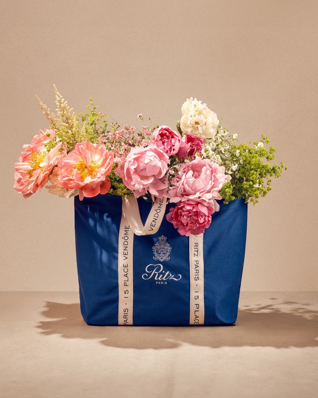 Still life of a blue Ritz Paris tote bag overflowing with a colorful variety of peony flowers in a studio setting with soft lighting.
