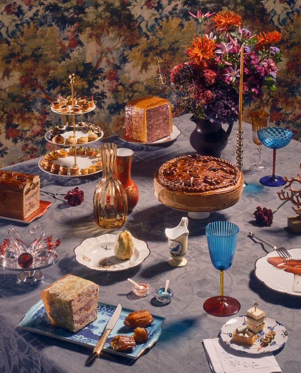 Elaborate table setting with cakes, pastries, flowers, glassware, and cutlery against a decorative floral wallpaper background.