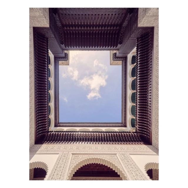 Low-angle architectural shot of a building's courtyard with white walls, arched doorways, and a wooden ceiling, looking up at the sky.