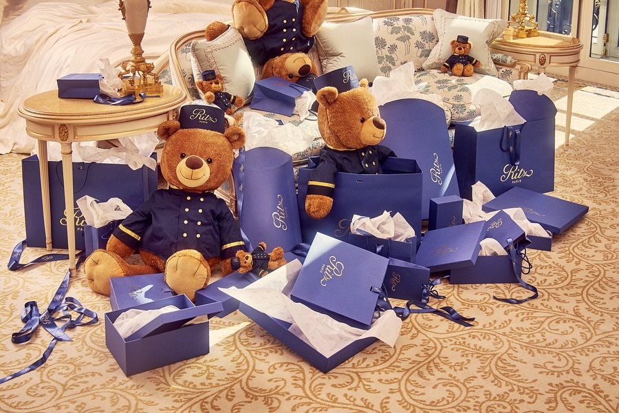 Teddy bears wearing uniforms are surrounded by blue shopping bags and boxes on a patterned carpet, creating a playful and luxurious scene.