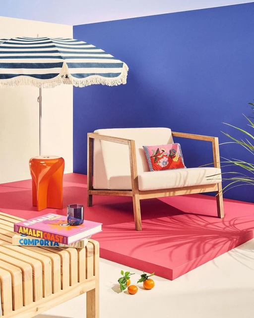 Interior setting features a cream-colored modern chair, a striped beach umbrella, a slatted bench, books, oranges, and vibrant color blocks.