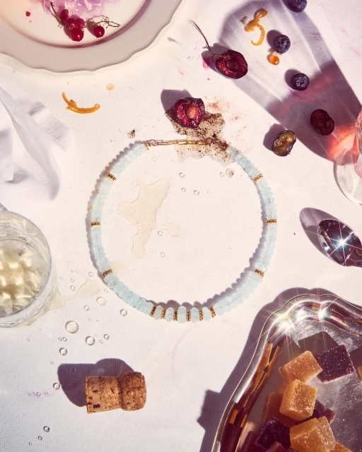 Overhead shot of an aquamarine beaded necklace, scattered berries, cork, and a silver tray of confectioneries against a white backdrop.