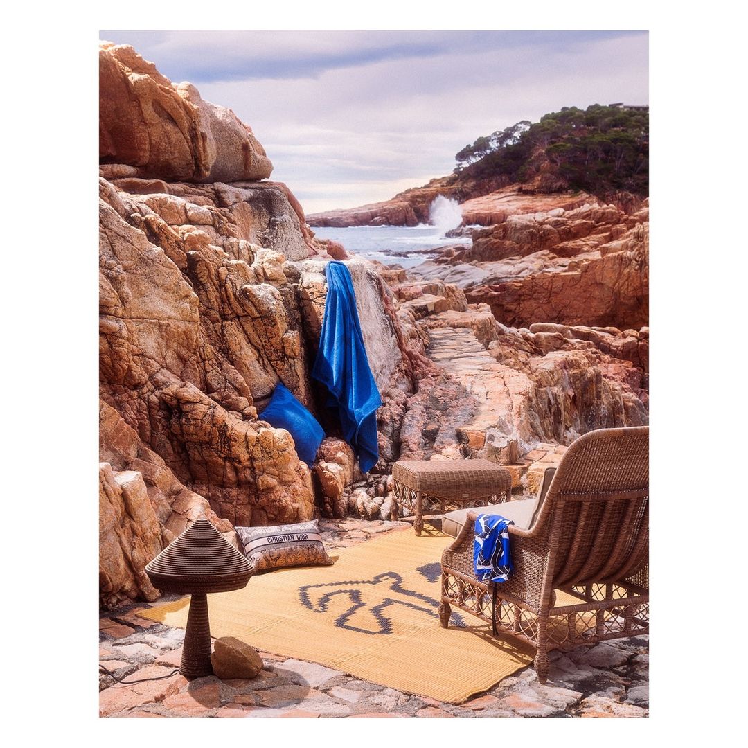 Coastal scene with a rocky beach, blue water, and wicker furniture set. A woven rug with an outline of a human figure sits on the ground.
