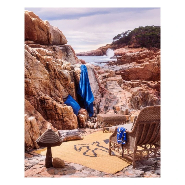 Coastal scene with a rocky beach, blue water, and wicker furniture set. A woven rug with an outline of a human figure sits on the ground.