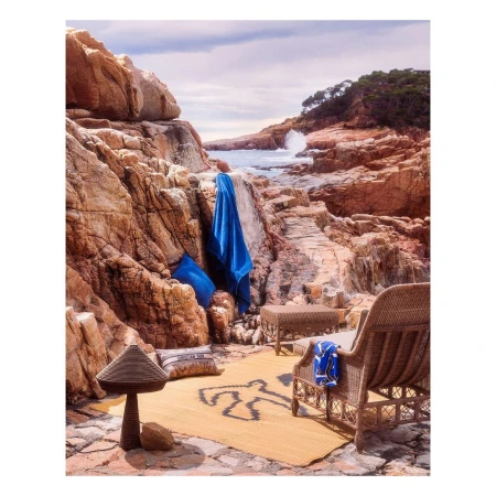 Coastal scene with a rocky beach, blue water, and wicker furniture set. A woven rug with an outline of a human figure sits on the ground.
