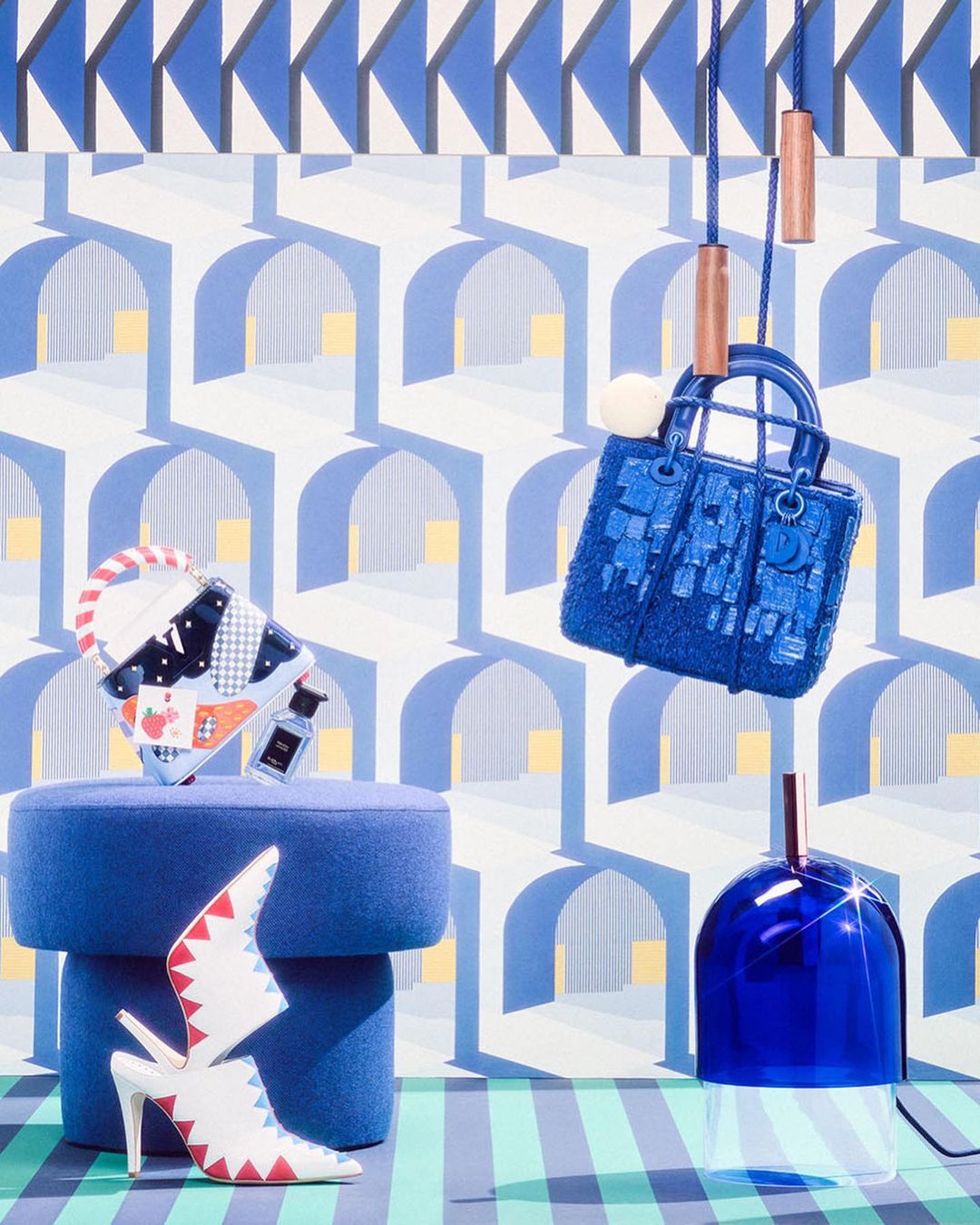 Eye-level shot of a blue purse, decorative shoes, and a blue glass container against a patterned backdrop. Studio product shot.