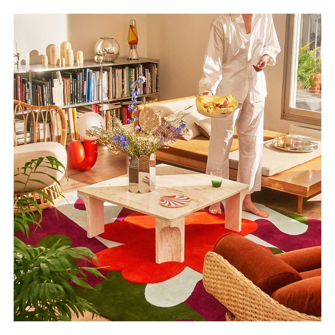 Person in white outfit holding plate with food near a coffee table on a colorful rug in a sunlit living room with books and plants.