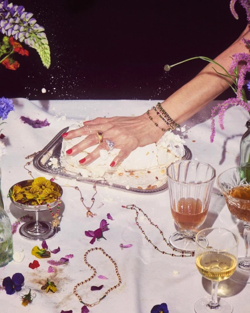 Close-up of a woman's hand smashing a cake on a table with champagne glasses, flowers, and jewelry, against a dark backdrop.
