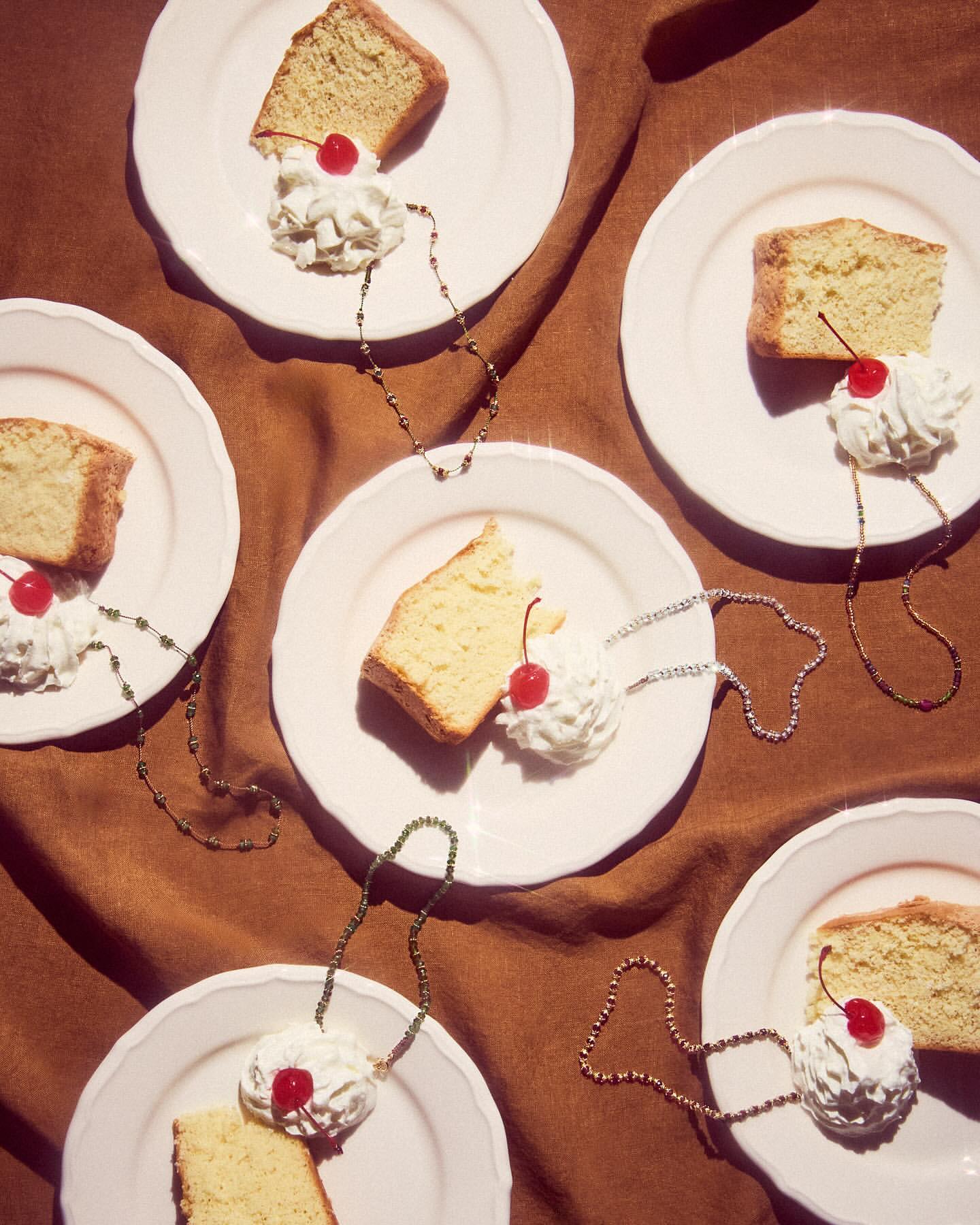 Overhead shot of multiple plates of pound cake topped with whipped cream and a cherry, each adorned with a beaded necklace on a brown cloth.