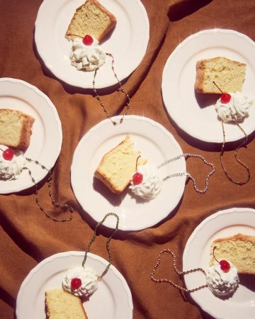Overhead shot of multiple plates of pound cake topped with whipped cream and a cherry, each adorned with a beaded necklace on a brown cloth.