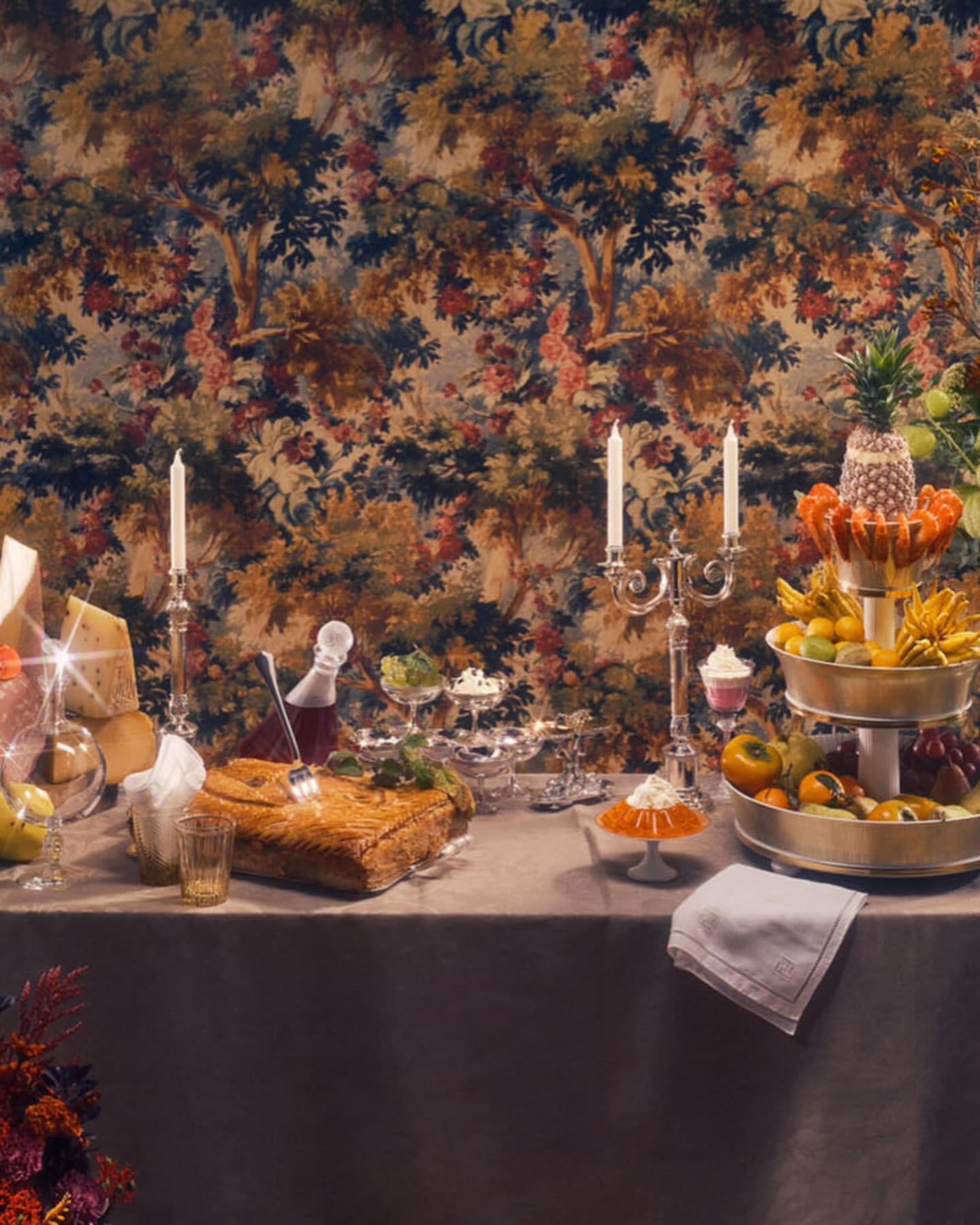 Table setting with assorted fruits, cheeses, and pastries, enhanced by decorative candles and a rich floral wallpaper backdrop.