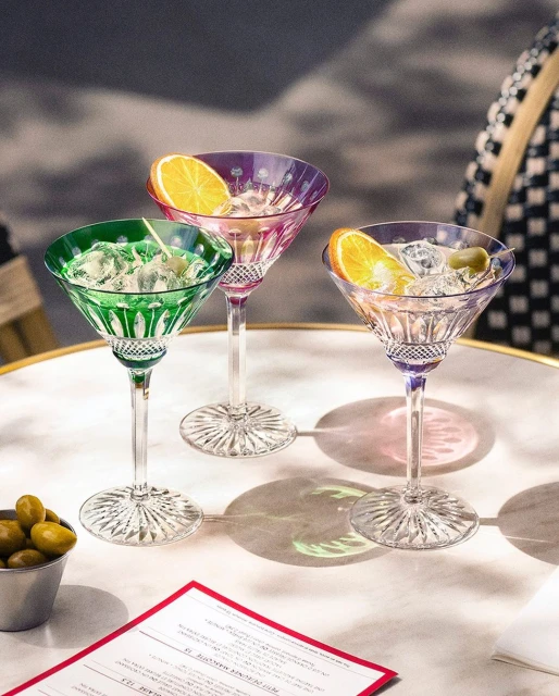 Three colorful crystal martini glasses with orange slice garnish stand on a table, casting shadows in a food and beverage setting.