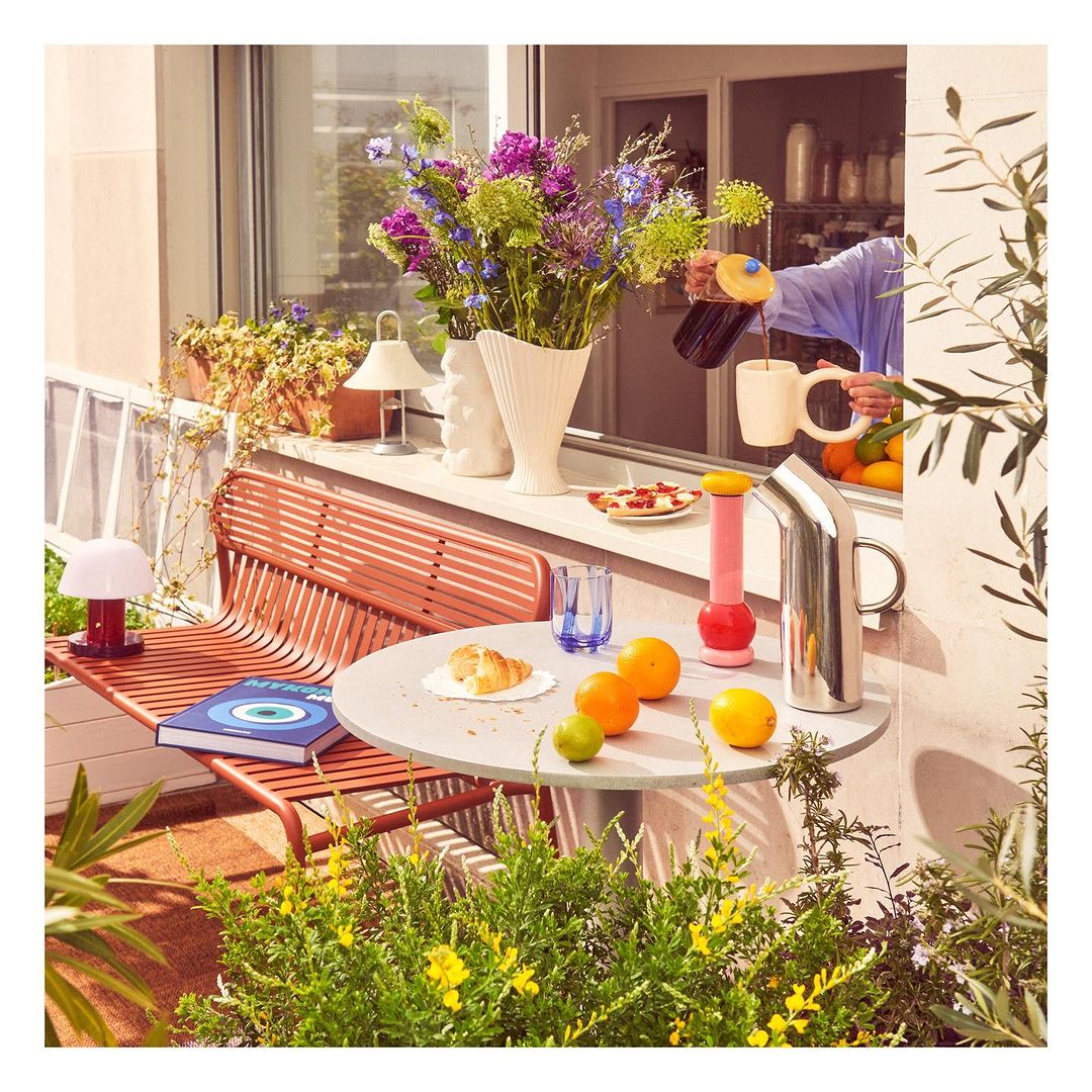 Person pouring dark beverage into a white mug on an outdoor table with fruit, flowers, and plants in a balcony setting.