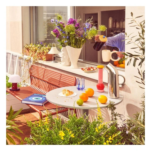 Person pouring dark beverage into a white mug on an outdoor table with fruit, flowers, and plants in a balcony setting.