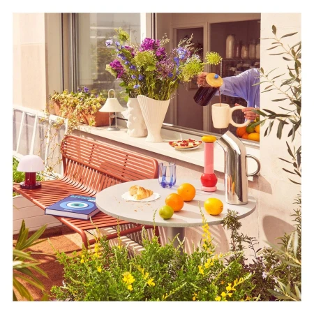 Person pouring dark beverage into a white mug on an outdoor table with fruit, flowers, and plants in a balcony setting.