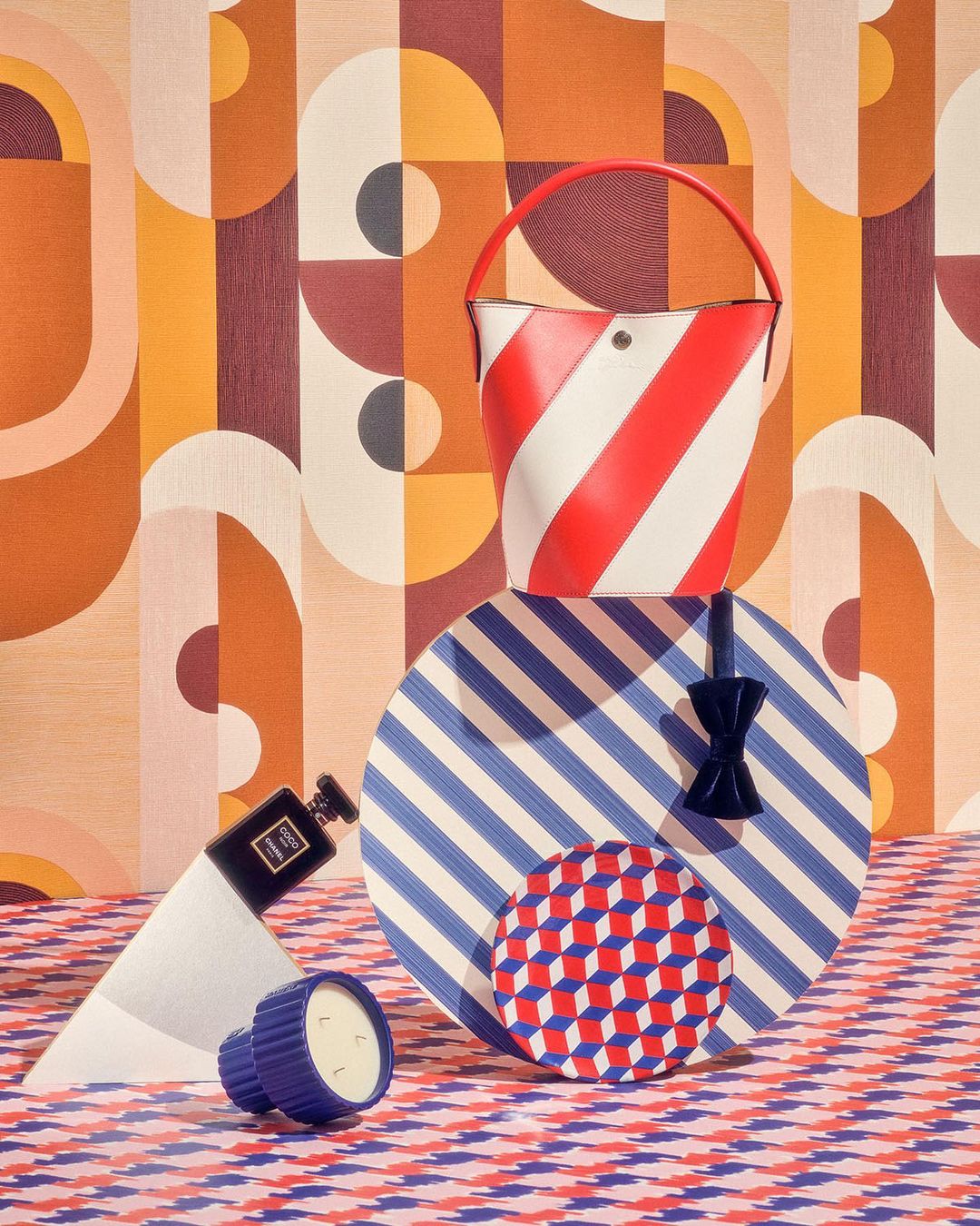 Still life featuring a red and white striped handbag, geometric patterned objects and surfaces, perfume, and candle on a table.
