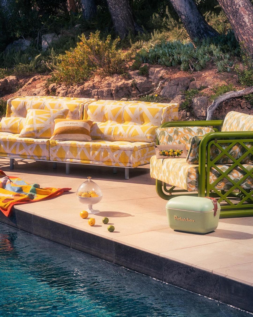 Outdoor patio with citrus-patterned sofa and chair next to a swimming pool, featuring lemons, limes, a cooler, and nearby greenery.