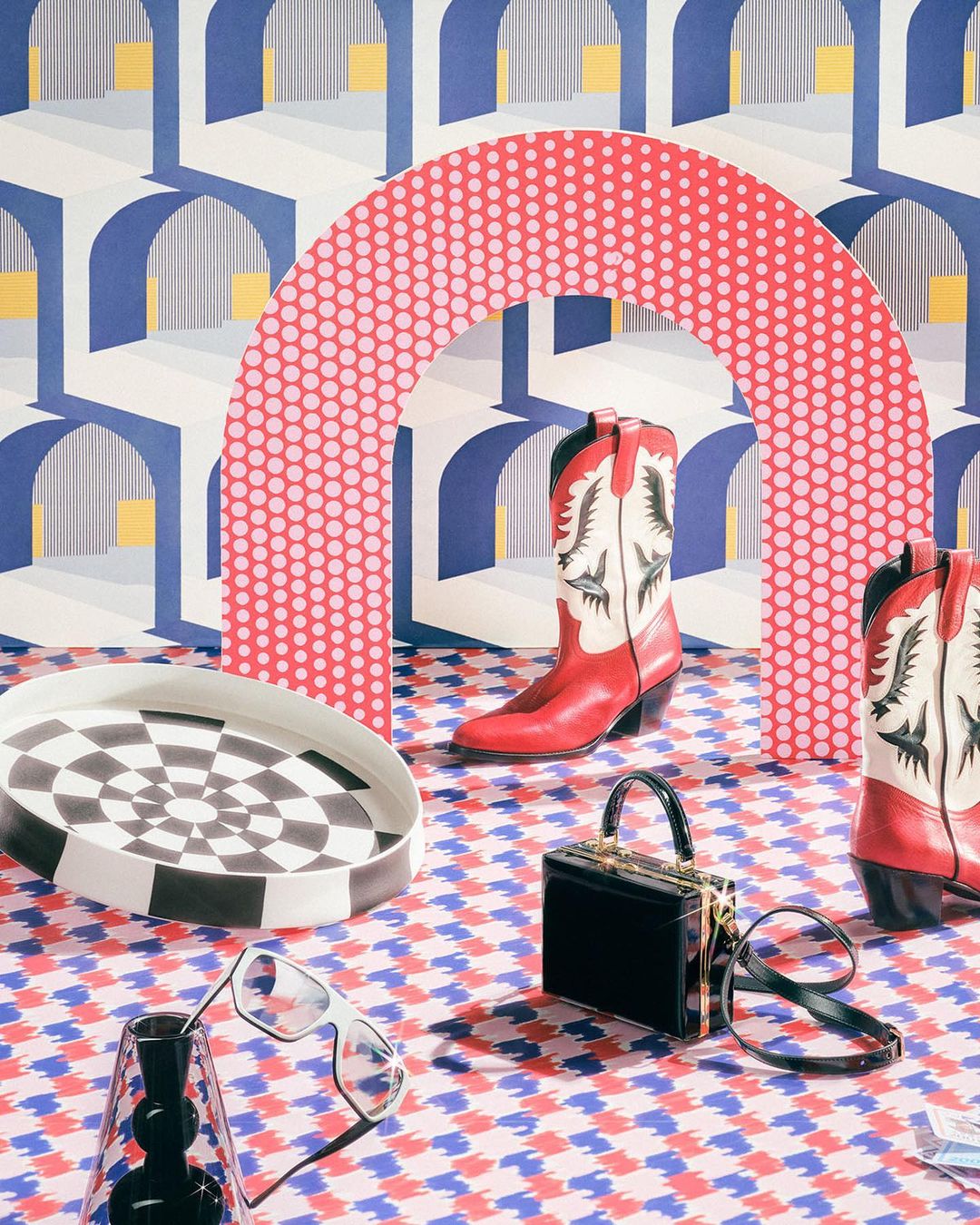 Still life shot of red and white cowboy boots with a black purse, eyeglasses, and checkerboard platter against a geometric backdrop.