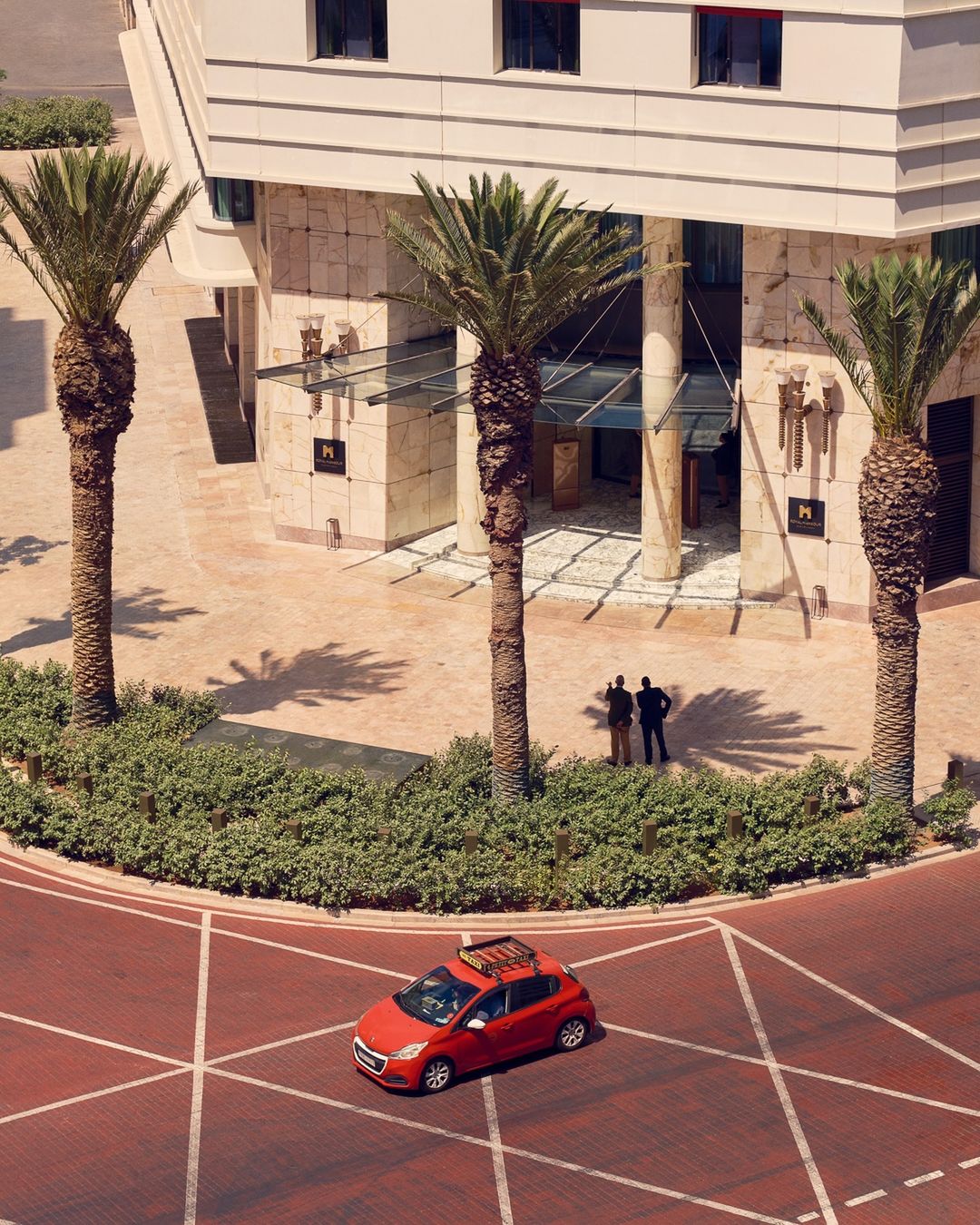 Red car parked on a brick road with palm trees and a building in the background, highlighting urban travel and architectural design.