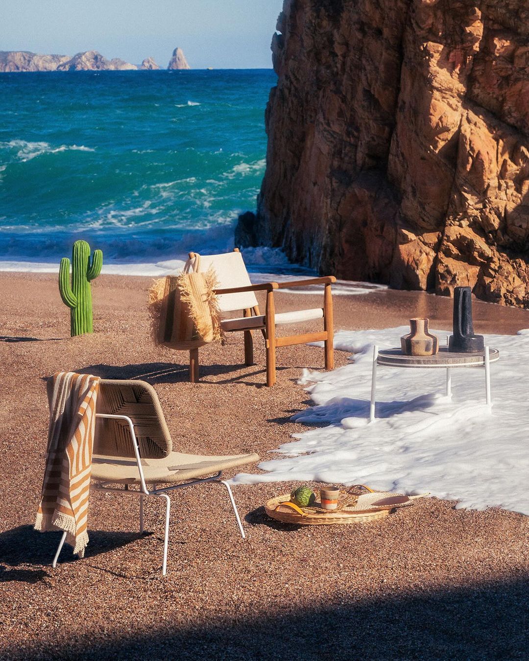 Coastal scene with two modern chairs, table with vases, and beach bag, set on sand near ocean waves and rocky cliffs under bright light.