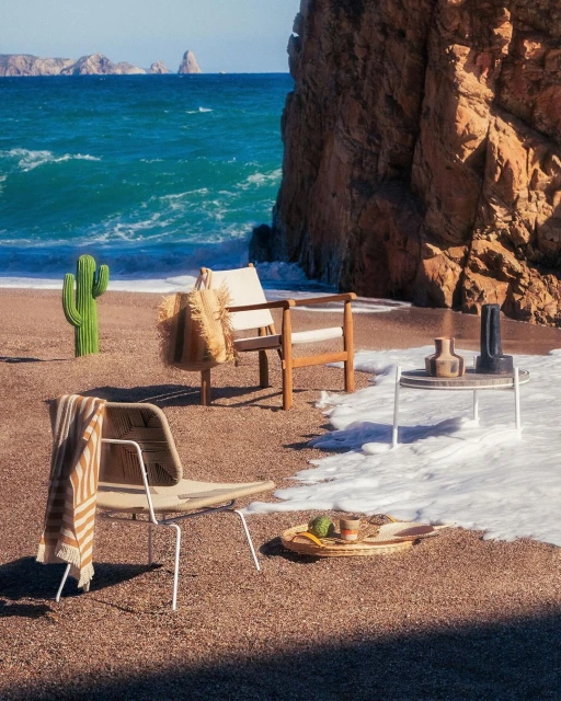 Coastal scene with two modern chairs, table with vases, and beach bag, set on sand near ocean waves and rocky cliffs under bright light.