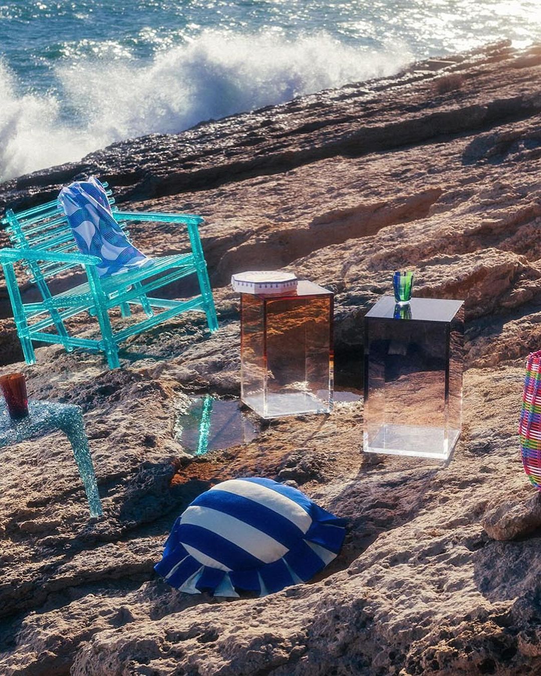 Outdoor product photoshoot on rocky beach with various furniture pieces, set against backdrop of crashing waves and blue ocean water.