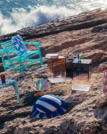 Outdoor product photoshoot on rocky beach with various furniture pieces, set against backdrop of crashing waves and blue ocean water.