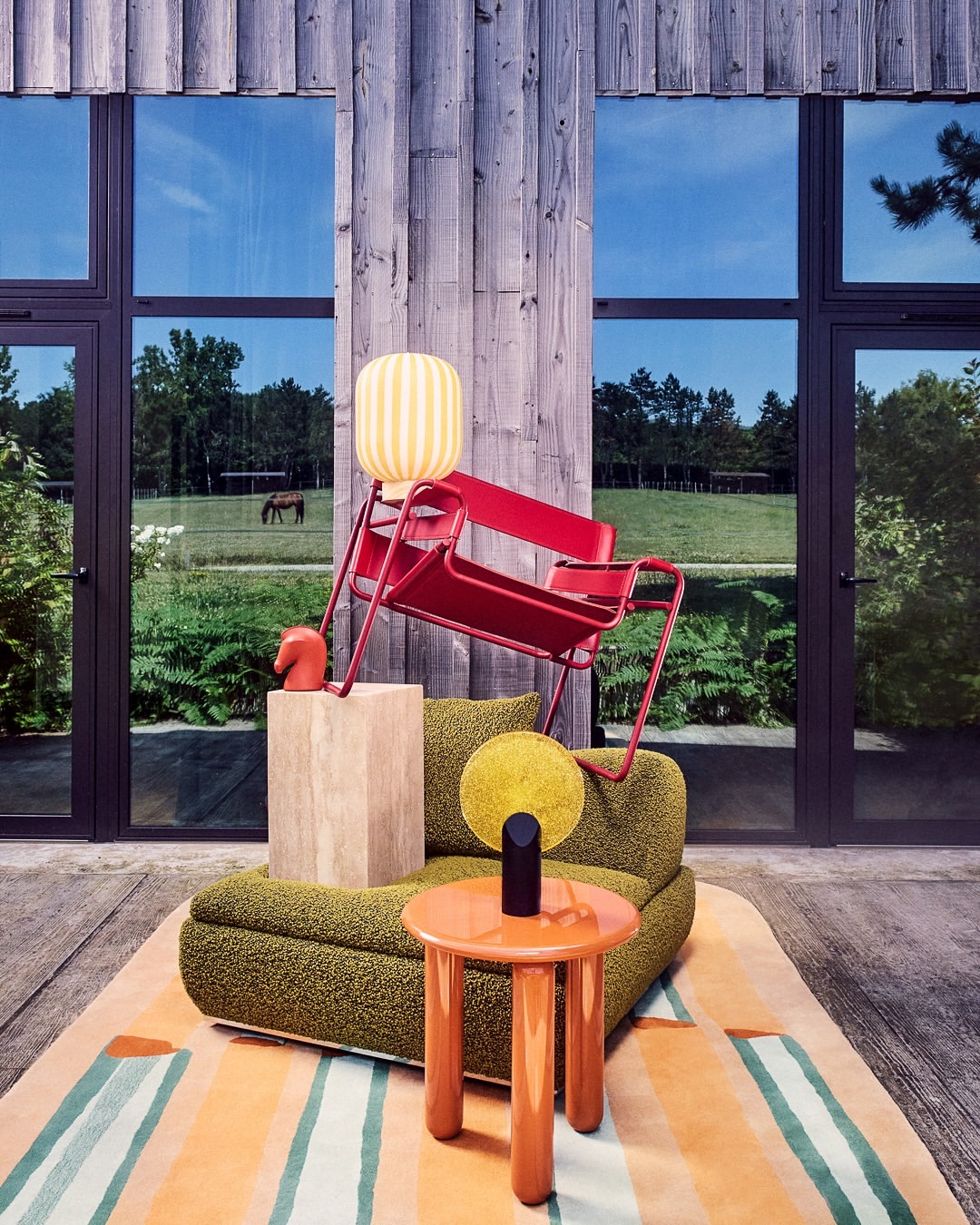 Indoor scene features a red chair, green sofa, and orange table on a striped rug, set against a wall with large windows and a pastoral view.