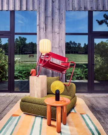 Indoor scene features a red chair, green sofa, and orange table on a striped rug, set against a wall with large windows and a pastoral view.