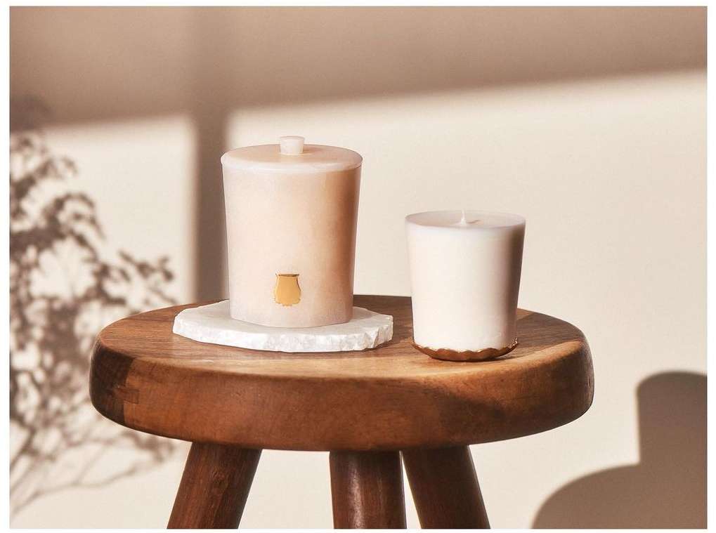 Two candles on a wooden stool against a neutral wall with plant shadows. One candle has a lid and a small decorative vase symbol.