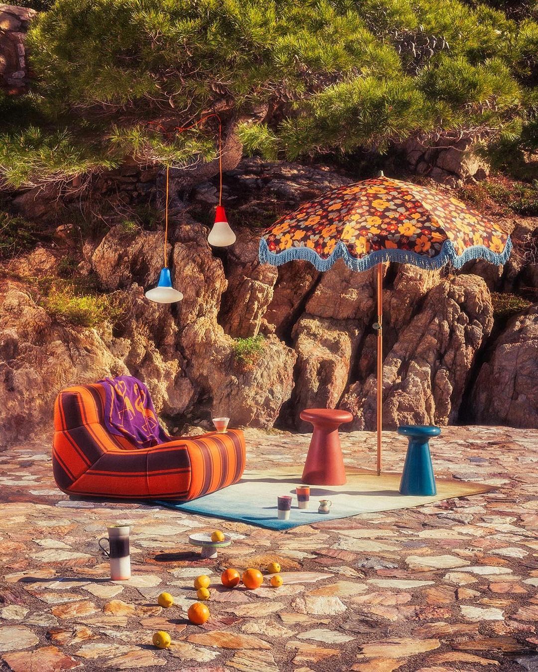 Outdoor lounge area with orange striped chair, umbrella, and colorful lights, set on a stone terrace amidst rocks and greenery.
